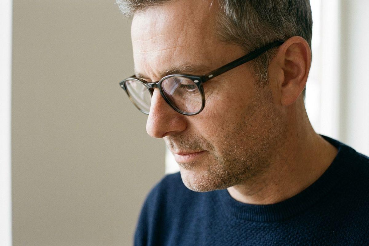 Close-up of an adult with glasses and short grey hair in a navy sweater, looking down with a serious expression beside a pale wall.
